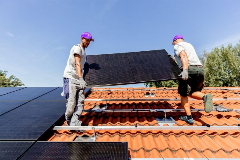 Zwei Monteure tragen ein PV-Modul auf ein mit roten Dachschindeln gedecktes Dach.