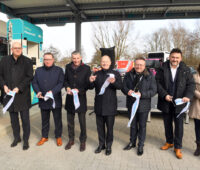 Eine Gruppe von Menschen in einer Reihe, die jeweils den Teil eines zerschnittenen Bandes in den Händen halten. Sie stehen vor einer Wasserstofftankstelle.