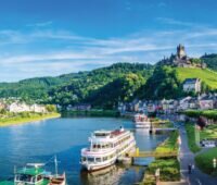 Die Mosel mit Hügeln und einer Burg im Hintergrund. Auf den Fluss sind Schiffe zu sehen, die an einer Anlegestelle liegen.