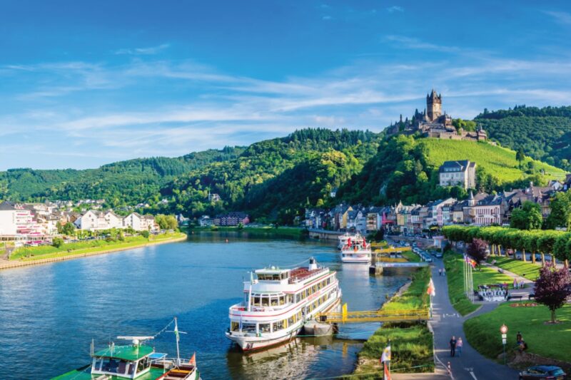 Die Mosel mit Hügeln und einer Burg im Hintergrund. Auf den Fluss sind Schiffe zu sehen, die an einer Anlegestelle liegen.