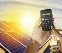 Taschenrechner wird von Händen hochgehalten. Im Hintergrund eine Photovoltaikanlage auf einem Dach.