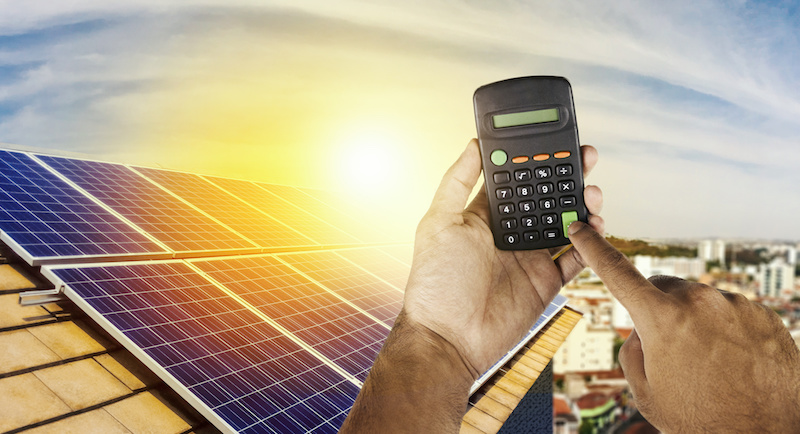 Taschenrechner wird von Händen hochgehalten. Im Hintergrund eine Photovoltaikanlage auf einem Dach.