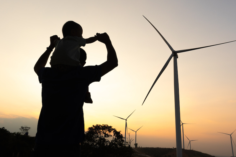 Im Gegenlicht: Ein Kind sitzt auf den Schultern eines größeren Menschen und blickt in Richtung Windkraftanlagen