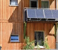 Eine Hausfassade mit Holzverkleidung, daran ein Balkon mit einer Steckersolaranlage mit 2 Photovoltaik-Modulen