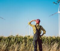 Frau in bäuerlicher Arbeitskleidung schaut in die Ferne auf einen Windpark - Symolbild für den Blick in die ungewisse Zukunft des EEG.