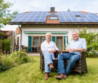 Ein Paar sitzt im Garten vor einem Einfamilienhaus mit Photovoltaikanlage auf dem Dach.