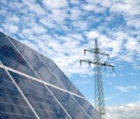 Ein Solarmodul im Vordergrund, dahinter ein Strommast unterwegs blauem Himmel mit Wolken.