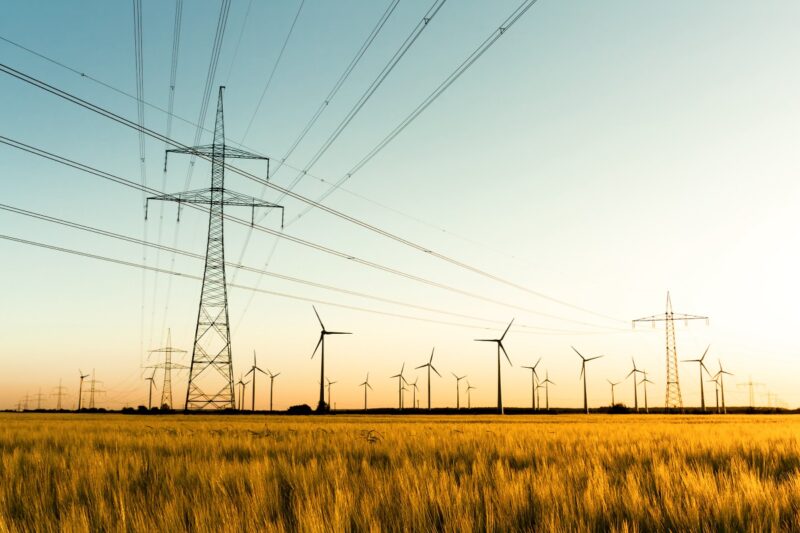 Stromnetz mit Feld und Windpark im Hintergrund bei Sonnenuntergang.