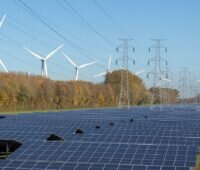 Im Vordergrund eine dunkel schimmernde Freiflächen-Photovoltaikanlage, dahinter Bäume, hinter denen wiederum die weißen Flügel von Windkraftanlagen sichtbar sind. In der Mitte und oben rechts im Bild sind Strommasten zu sehen. Ein Symbolbild für den Artikel, in dem es um neue Regeln für Stromnetzentgelte geht.