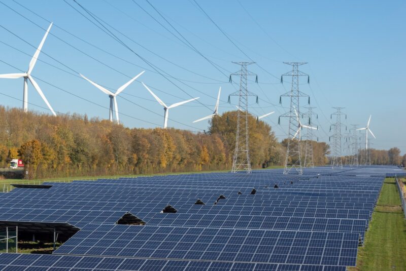 Im Vordergrund eine dunkel schimmernde Freiflächen-Photovoltaikanlage, dahinter Bäume, hinter denen wiederum die weißen Flügel von Windkraftanlagen sichtbar sind. In der Mitte und oben rechts im Bild sind Strommasten zu sehen. Ein Symbolbild für den Artikel, in dem es um neue Regeln für Stromnetzentgelte geht.