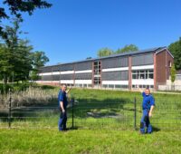 Zwei Männer im Blaumann an einem Zaun, hinter dem eine Schule mit PV-Anlage auf dem Dach steht.