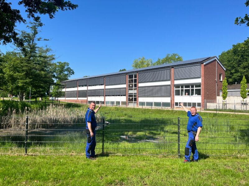 Zwei Männer im Blaumann an einem Zaun, hinter dem eine Schule mit PV-Anlage auf dem Dach steht.