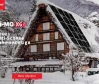 Hi-MO X6 von Longi mit neuem Anti-Schnee Rahmendesign. Zu sehen ist ein altes schneebedecktes Holzhaus mit spitzem Dach in einer bergigen Winterlandsschaft. Das Dach ist mit Photovoltaikmodulen belegt, welche schneefrei sind.