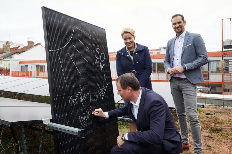 Solarmodul mit gezeichneter Sonne und Unterschriften, ein Mann unterschreibt, eine Frau und ein Mann stehen daneben.