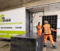 ein grün-weißer Container mit geöffneter Klappe, ein Stapel Photovoltaik-Module und zwei Menschen mit orangefarbenen Anzügen: der mobile Teststand des Recycling-Anbieters Buhck