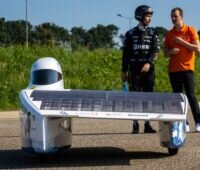 Zwei Menschen stehen hinter einem Solarauto, ein schmaler flacher Flitzer mit einem seitlichen Flügel mit Solarzellen.