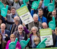 Gruppe von Menschen aus der Vogelperspektive demonstriert mit Pappschildern und überdimensionalen Schaumstoffhänden für Bürgerenergie, u.a. Energy Sharing.