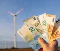 Hand mit Geldscheinen, im Hintergrund Windrad - Symbolbild für Klimageld und Energiewende.