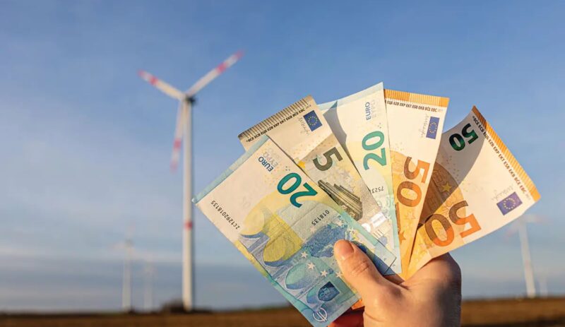 Hand mit Geldscheinen, im Hintergrund Windrad - Symbolbild für Klimageld und Energiewende.