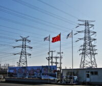 Strommasten und Leitungen vor blauem Himmel. Mittig eine wehende chinesische Fahne.