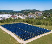Eine solarthermische Kollektoranlage auf grüner Wiese vor einer Fabrik und Hügeln im Hintergrund.