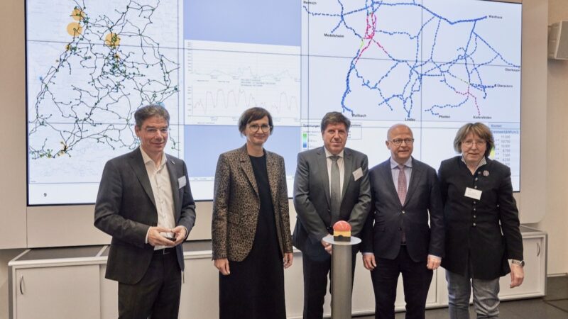 Drei Männer und zwei Frauen hinter rotem Knopf und vor Karte des Energiesystems - Start für den digitalen Zwilling