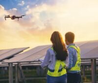 Ein Mann und eine Frau vor einer Photovoltaik-Anlage, darüber fliegt eine Drohne - Thermografie von Prüfhelden.