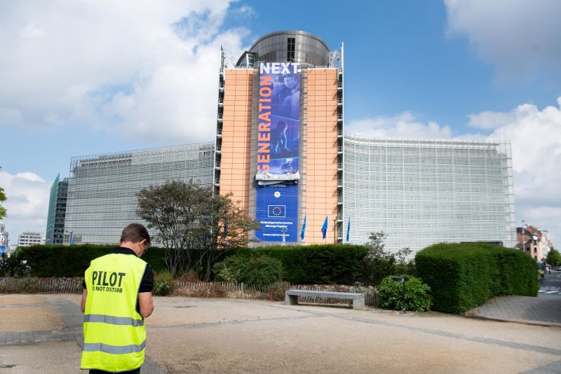 Am Sitz der EU-Kommission in Brüssel hängt ein Plakat mit der Aufschrift EU NExt Generation, im Vordergrund in Arbeiter in Sicherweste mit dem Rücken zum Betrachter auf den Boden schauend.