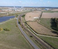 Luftbild zeigt Wiesen, Felder und eine Landstrasse - im Hintergrund eine Industrieansiedlung.