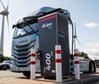 Lkw-Zugmaschine hinter Ladepunkt, im Hintergrund ein Windrad- Symbol für Förderung für elektrische Nutzfahrzeuge