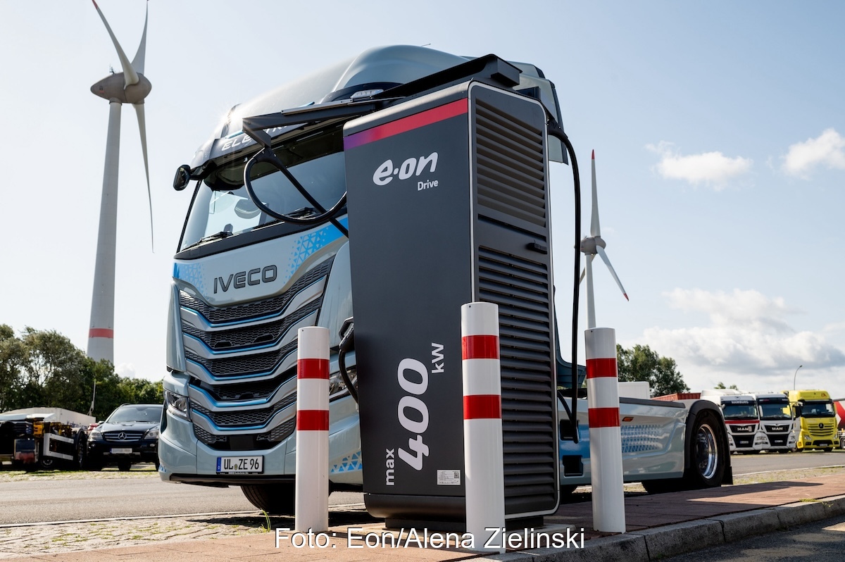 Lkw-Zugmaschine hinter Ladepunkt, im Hintergrund ein Windrad- Symbol für Förderung für elektrische Nutzfahrzeuge