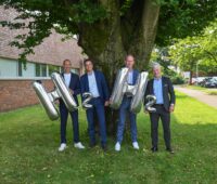 Vier Männer in Anzügen tragen Luftballons mit H und 2.