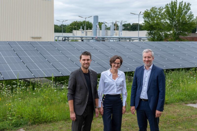 Drei freundlich blickende Personen vor einer PV-Anlage und einer Industrieinstallation.