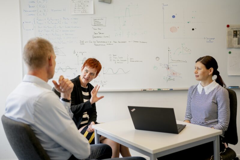 Ein Mann und eine Frau gestikulierend im Gespräch, eine junge Frau sitzt hinter einem Computer - Forschungsgruppe für Energiewende-Kommunikation.