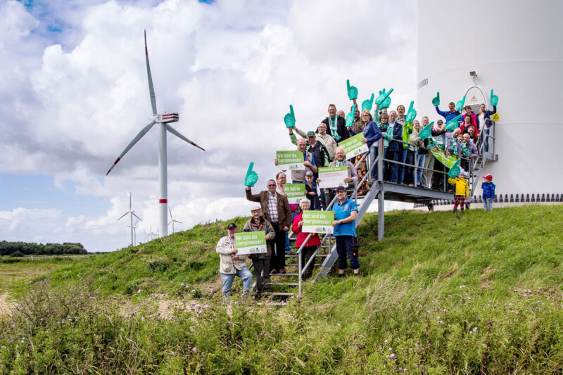 Menschengruppe zeigt am Fuß einer WIndenergieanlage Plakate und ähnliches, um für Bürgerenergie und Energy Sharing zu demonstrieren.