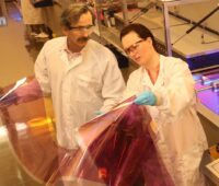 Im Bild sind zwei Wissenschaftler:innen, die Solarzellen aus dem Drucker in den Händen halten, die für Multi-Benefit-PV geeignet sind.