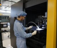 Ein Laborant hält eine Solarzelle vor einer Maschine in der Hand.