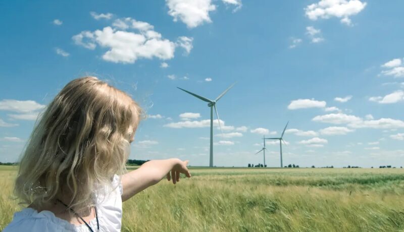 Ein Kind deutet über ein Weizenfeld auf Windenergie-Anlagen - Symbolbild für Genehmigungen.