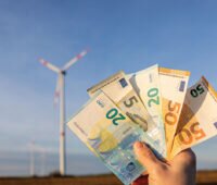 Hand hält Geldscheine vor Windpark in die Höche - Symbol für finanzielle Beteiligung von Kommunen