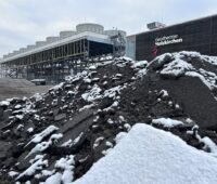 Dunkler Erdhaufen mit etwas Schnee, im Hintergrund ein Gebäude mit der Aufschrift "Geothermie Holzkirchen"