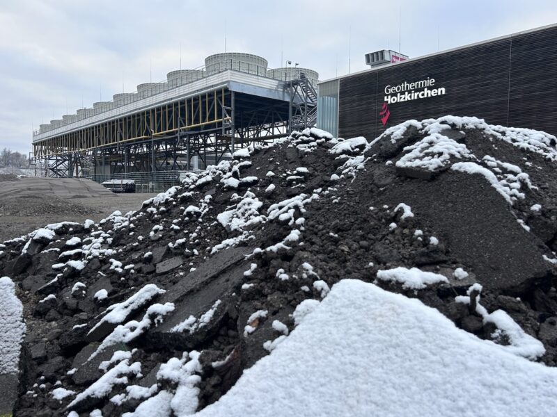 Dunkler Erdhaufen mit etwas Schnee, im Hintergrund ein Gebäude mit der Aufschrift "Geothermie Holzkirchen"