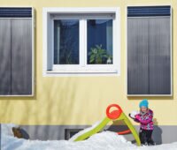 Solar-Luftkollektoren an einer gelben Hauswand montiert. Davor spielt ein Kind im Schnee.