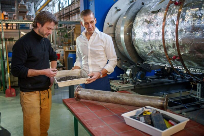 Zwei Männer in einem Industriegebaäude mit Werkstoffen und der schwarzen Masse alter Batterien.