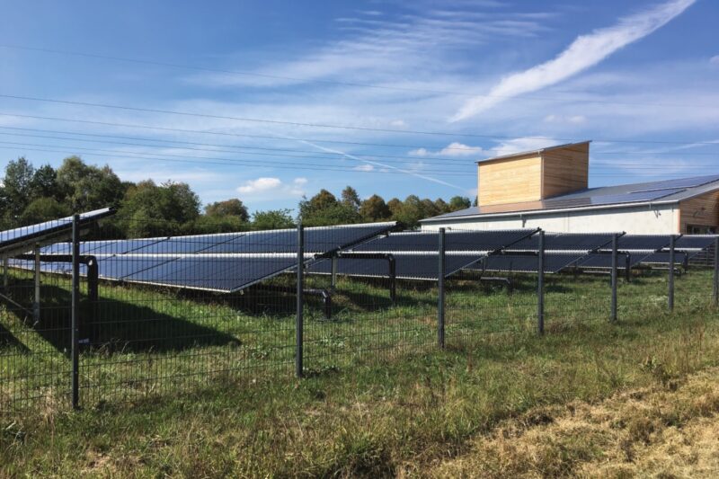 Hinter einem Zaun ist eine größere Solarthermieanlage mit Röhrenkollekoren installiert. Im Vordergrund eine grüne Wiese, im Hintergrund blauer Himmel. Am rechten Rang ragt ein kleiner Holzturm emport - er gehört zu einer Holzhackschnitzelanlage
