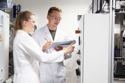 Ein Mann und eine Frau in weißen Laborkitteln mit einer Probe - Forschungsprojekt für Wasserstoff aus Abwasser.