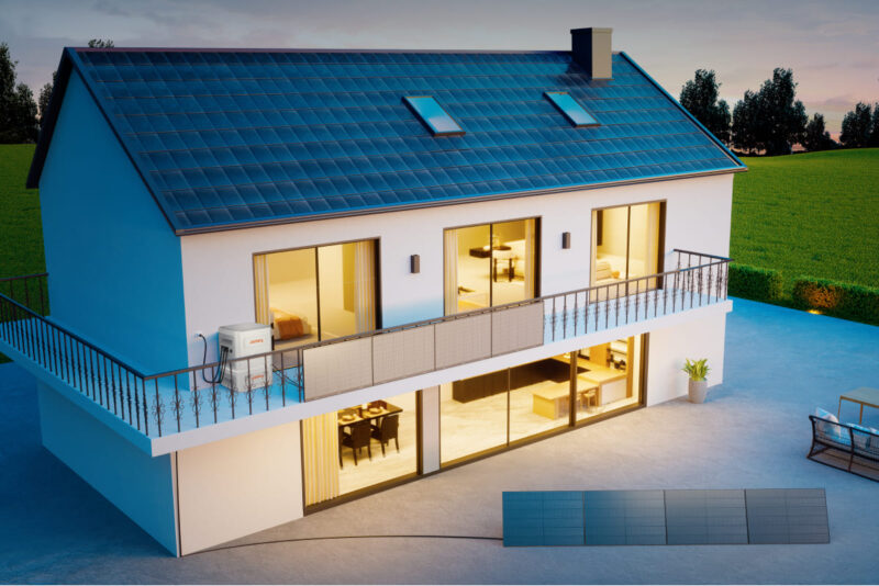 Zu sehen ist ein modernes von innen beleuchtets Einfamilienhaus mit einem mobillen Speicher von Jackery auf dem Balkon und angeschlossenen PV-Modulen, die auf der Terasse aufgestellt sind.