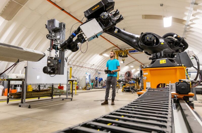 Mann mit Kontroll-Panel i einer Werkhalle mit Roboter und einem Windturbinenblatt.