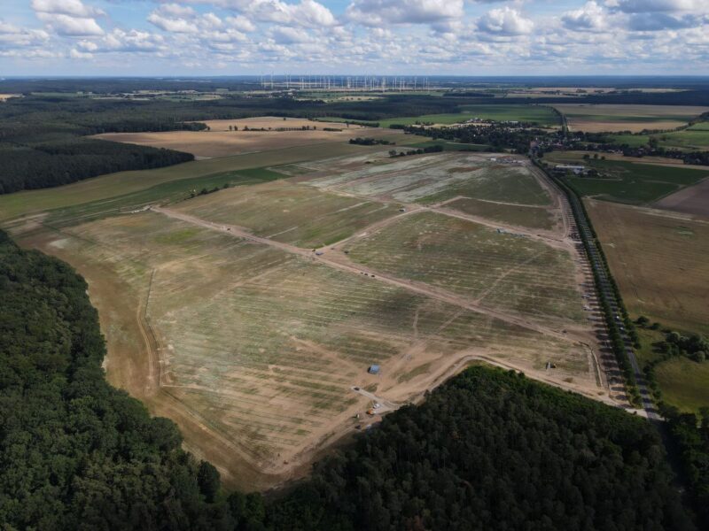 Luftbild zeigt eine vorbereitete Fläche zum Bau eines großen Freiflächen-Solarparks umgeben von Wäldchen und Feldern.