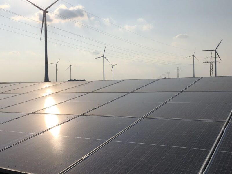 Photovoltaik mit refelktiernder Sonne im Vordergrund, dahinter Windkraftanlagen.