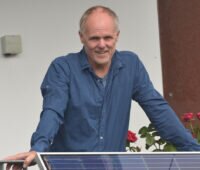 Jörg Lange, ein Mann im "besten Alter" stützt sich auf ein Balkongeländer, an dem ein Photovoltaikmodul als Balkonkraftwerk befestigt ist.